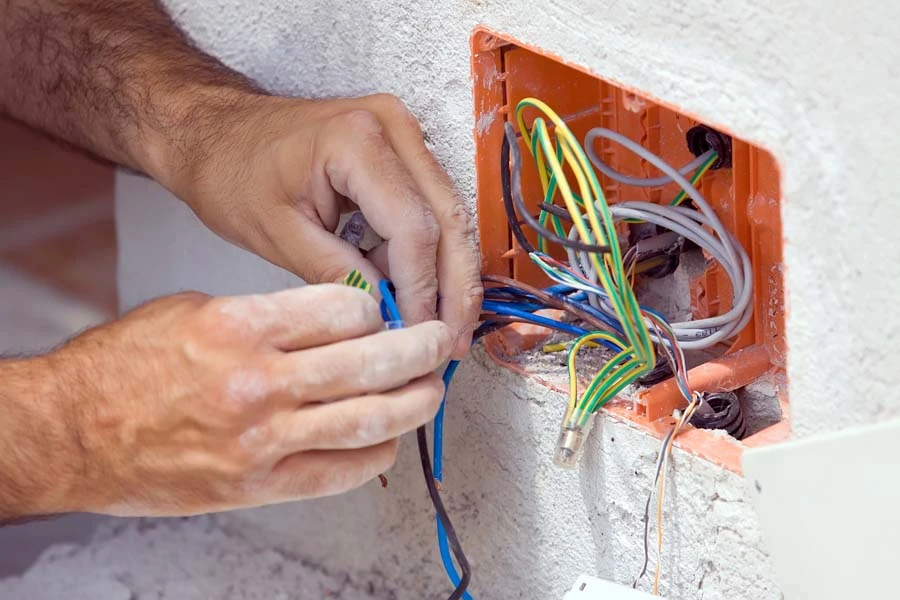 Électricien travaillant sur installation électrique murale, manipulant des câbles multicolores sortant d'un boîtier orange encastré dans mur blanc