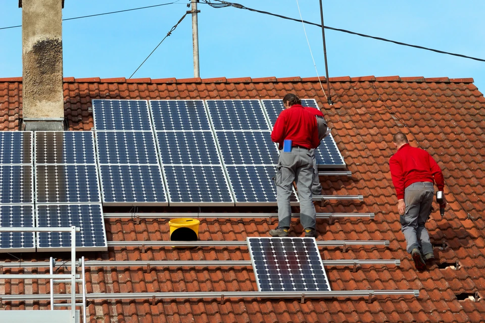 Deux techniciens en uniformes rouges installent des panneaux solaires photovoltaïques sur une toiture en tuiles rouges d'une maison résidentielle
